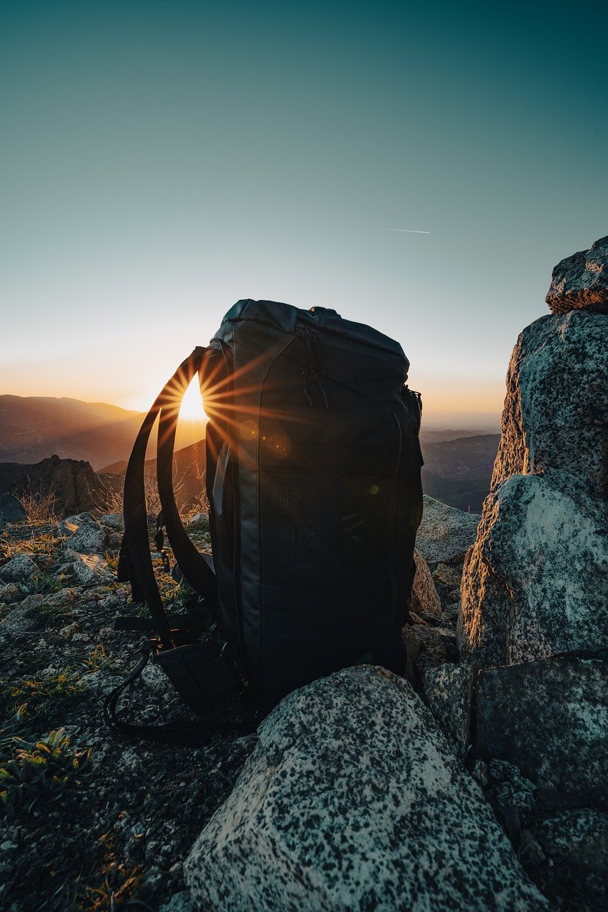 Rucksack auf felsigem Berggipfel bei Sonnenaufgang, mit Sonne im Hintergrund und weiter Landschaft im Dunst.
