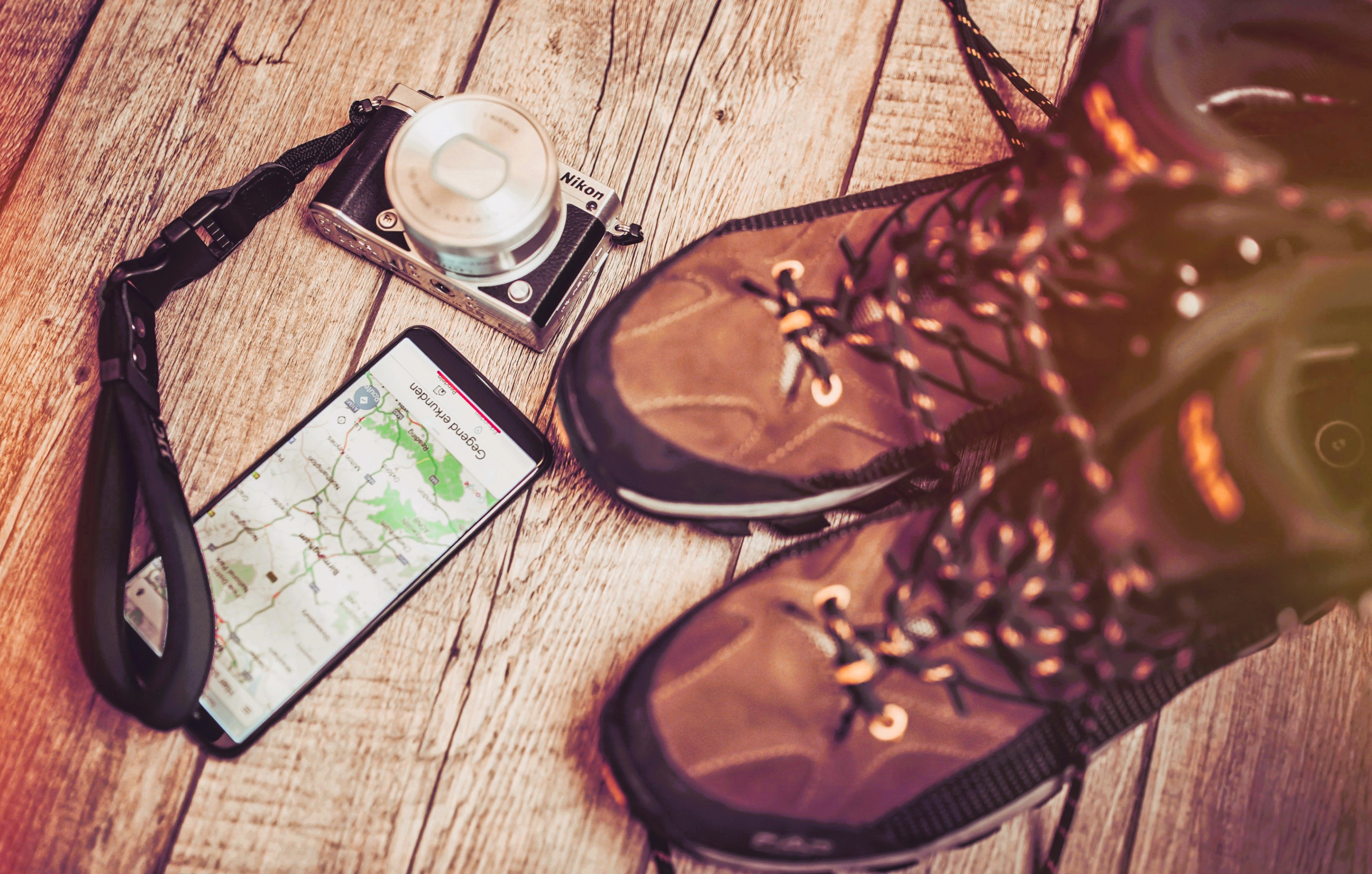 Wanderstiefel, Kamera und Smartphone mit geöffneter Karten-App auf einem Holzboden arrangiert, als Vorbereitung für eine Reise oder Wanderung.