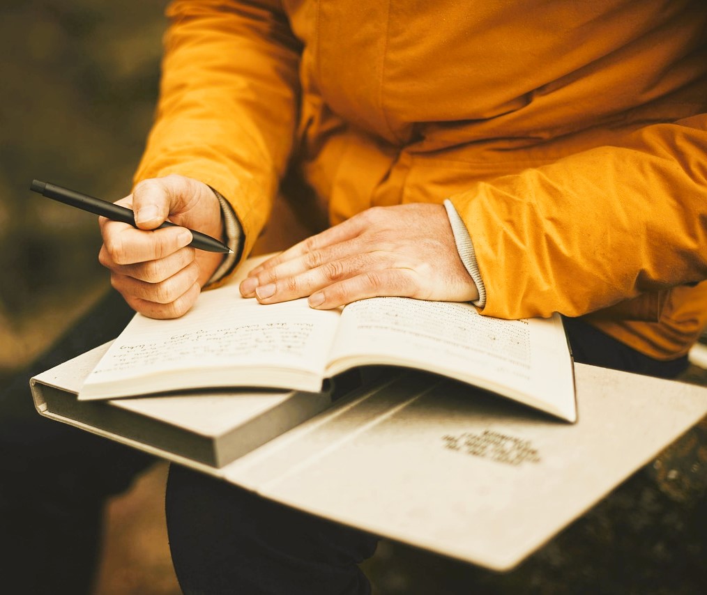 Person in orangefarbener Jacke schreibt mit einem Stift in ein aufgeschlagenes Notizbuch im Freien.